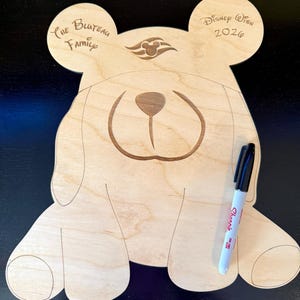 May include: A light-colored wooden cutout shaped like a bear with rounded ears, featuring engraved details including a nose, mouth, and the text "The Bluteau Family" and "Disney Wish 2026". A black Sharpie marker is placed on the right side of the bear.