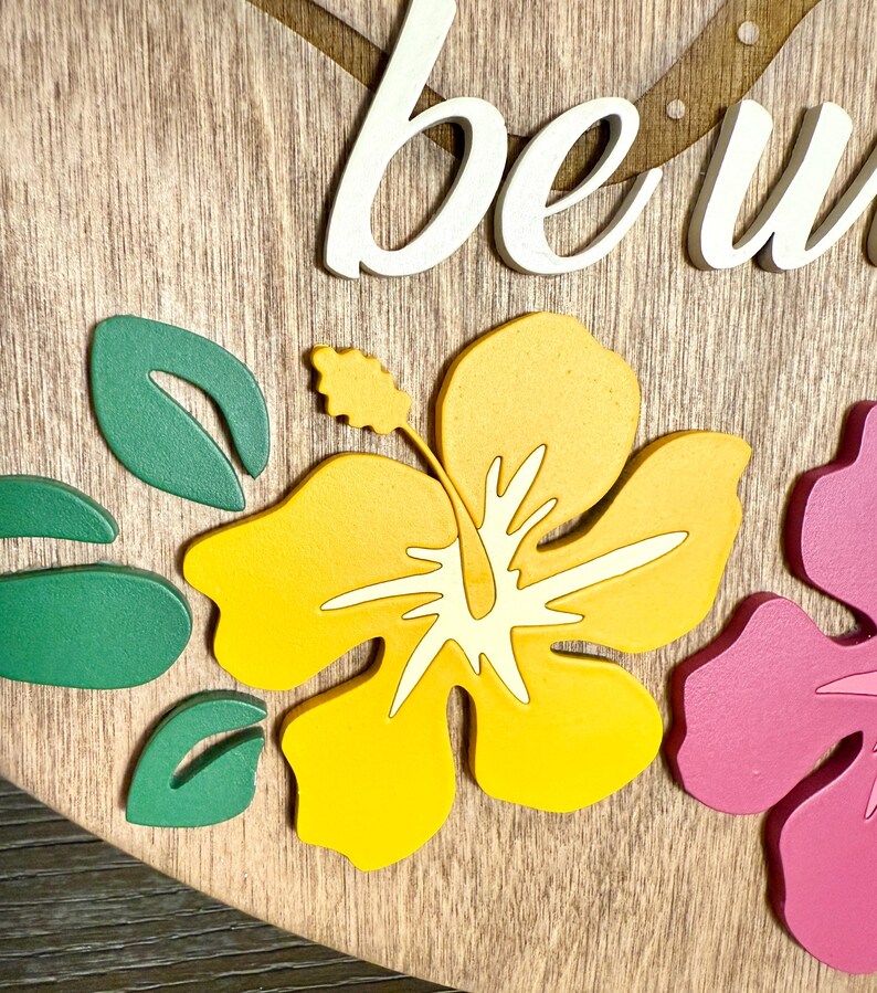 May include: Wooden sign with the word "be" in white script, featuring a yellow hibiscus flower with white accents, green leaves, and a pink flower. The background is a light brown wood grain.