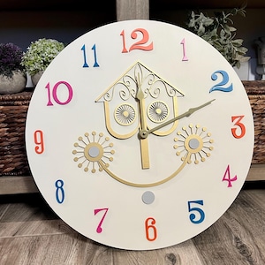 May include: A white round clock with gold accents and colorful numbers. The clock face features a stylized house with two round windows and a gold clock hand.