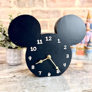 May include: A black wooden clock in the shape of Mickey Mouse's head. The clock face has white numbers and gold-colored hands. The clock is sitting on a marble surface.