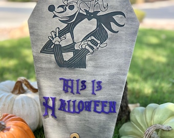 May include: A gray wooden coffin-shaped sign featuring a black illustration of Jack Skellington and Zero from The Nightmare Before Christmas. The sign reads "This Is Halloween" in purple letters, with an orange pumpkin at the bottom. Pumpkins are in the background.