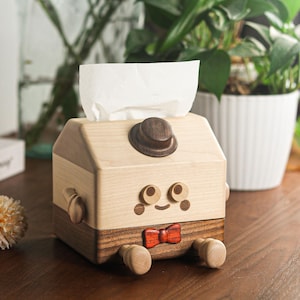 May include: Wooden tissue box with a whimsical design. The box is light-colored with a dark brown base and a small hat on top. It features a cartoon face with a red bow tie and wooden appendages.