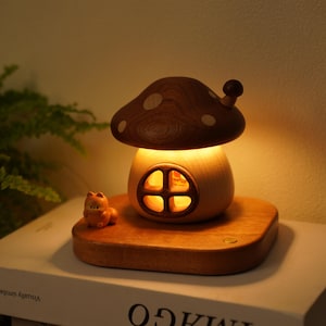 May include: A wooden mushroom-shaped night light with a brown cap and white spots. The light has a round window and sits on a wooden base. A small figurine is placed on the base. The light is turned on, emitting a warm glow.