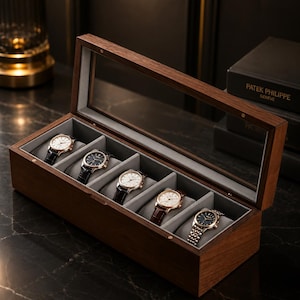 Puede incluir: Una caja de madera oscura para relojes con tapa de cristal, que muestra seis relojes de pulsera. Los relojes tienen esferas doradas y plateadas y diferentes estilos de correas, sobre un interior gris. La caja es rectangular.