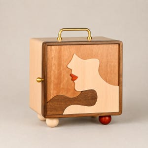 Pode incluir: Uma caixa de armazenamento quadrada de madeira com uma incrustação de marchetaria de um perfil facial no painel frontal. A caixa tem uma alça de metal dourado no topo, um pequeno botão dourado na lateral e quatro pés esféricos de madeira clara e vermelha escura.
