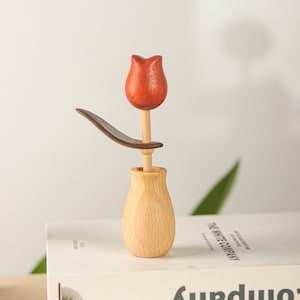 Può includere: Una scultura di tulipano in legno con un fiore rosso, una foglia marrone e un vaso di legno chiaro. Il tulipano è esposto su un libro con il testo "THE WHITE COMPANY". È un oggetto decorativo.