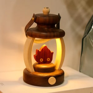 May include: A wooden lantern with a dark brown top and base, light wood frame, and a red flame-shaped figure inside. The lantern has a leather handle and a small wooden knob on top. The interior glows with a warm light.