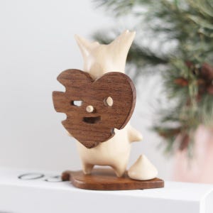 May include: A wooden figurine of a cartoon character holding a heart-shaped cutout with a smiling face. The figurine is standing on a wooden base.