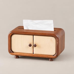 Puede incluir: Un dispensador de pañuelos rectangular de madera con un marco marrón oscuro y dos puertas estilo armario de color beige claro con pequeños pomos redondos. La parte superior tiene una ranura horizontal de la que sobresale un pañuelo blanco.