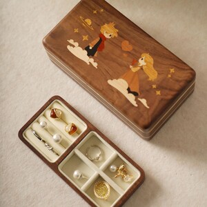 May include: A wooden jewelry box with a hinged lid, featuring an inlaid design of two figures on clouds, stars, and a planet. The open box reveals compartments with rings, earrings, and other jewelry. The box is made of dark wood.