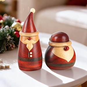 Può includere: Due statuette di Babbo Natale in legno. Una è alta e snella, l'altra è rotonda. Entrambe sono principalmente rosse con accenti in legno chiaro per la barba, il bordo del cappello e il viso. Il Babbo Natale alto ha una piccola palla di legno sulla cima del suo cappello.