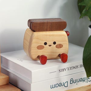 Puede incluir: Un juguete de madera con forma de tostada con una cara sonriente, mejillas sonrosadas y pies rojos. La parte superior de la tostada es de color marrón oscuro. El juguete está sobre una pila de libros blancos.