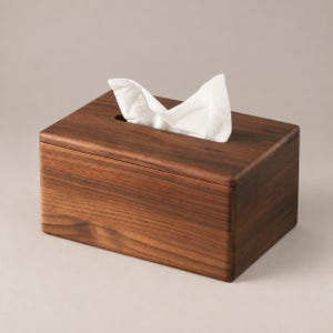 Puede incluir: Una caja rectangular de madera de color marrón oscuro con un acabado liso. Un pañuelo de papel blanco asoma a través de la abertura rectangular estrecha y centrada en la parte superior de la caja, creando un aspecto suave y plegado.