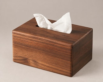 May include: A rectangular dark brown wooden tissue box cover with a smooth finish. A single white paper tissue is pulled through the narrow, centered rectangular opening on the top surface of the box, creating a soft, folded appearance.