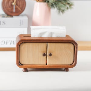 May include: A wooden tissue box holder with a cabinet design. The box has two doors with brown knobs and a slot for tissues on top. The box is made of light and dark wood.