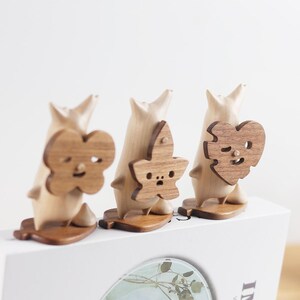 May include: Three wooden figurines with different facial expressions and leaf-shaped designs. The figurines are standing on a white surface.