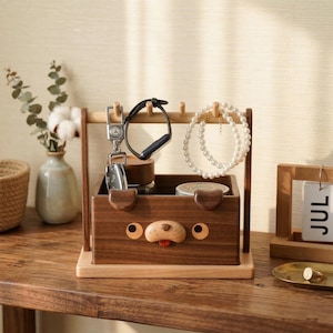 May include: A wooden desk organizer shaped like a dog, with a light brown base and a darker brown box. It features a key holder, watch holder, and a place to hang jewelry. The dog's face has a red tongue and round eyes.