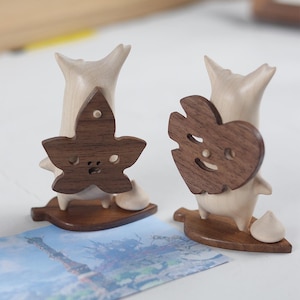 May include: Two wooden figurines with a leaf design on their backs. The figurines are made of light wood and have a dark wood leaf design. The leaf designs are different, one is a heart shape and the other is a maple leaf.