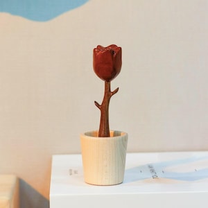 Può includere: Una scultura in legno di una rosa rossa in un piccolo vaso di legno di colore chiaro. La rosa ha un aspetto dettagliato e strutturato, con uno stelo marrone e una finitura in legno naturale sul vaso. È un oggetto decorativo.