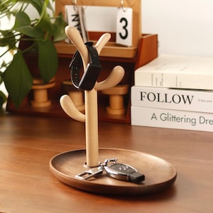 Puede incluir: Un organizador de madera con un soporte en forma de árbol y una bandeja redonda. El soporte tiene cuatro ramas para colgar objetos, y la bandeja es perfecta para guardar llaves, joyas u otros objetos pequeños. Un rastreador de fitness negro está colgado en una de las ramas, y una llave de coche está en la bandeja.