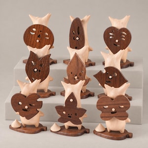Peut inclure: Une collection de neuf petites figurines en bois, chacune composée d'une base claire en forme de créature debout tenant un masque en bois brun foncé avec des traits faciaux découpés. Les masques varient en forme, incluant cercles, cœurs et étoiles.