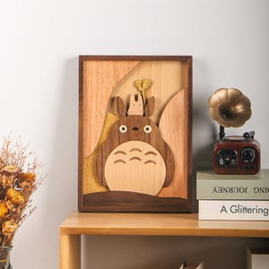 Puede incluir: Una pieza de arte mural rectangular de madera con una representación tallada y en capas de una criatura grande, marrón y de cuerpo redondo con orejas puntiagudas y una criatura más pequeña y clara sobre su cabeza, frente a árboles de madera.