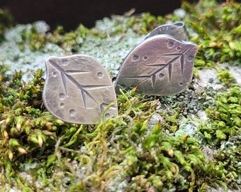 Puede incluir: Un par de pendientes de plata en forma de hoja con una superficie texturizada y un diseño delicado. Los pendientes se muestran sobre un lecho de musgo verde.