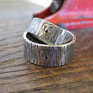 May include: Two silver wedding bands with a hammered texture. The bands are stacked on top of each other and feature a heart with the initials "TK" engraved inside.