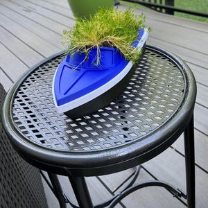 May include: A blue, white, and black boat-shaped planter filled with green plants. The planter sits on a black metal table with a patterned surface. The table has a round top and three legs. The background includes a green pot and wooden decking.