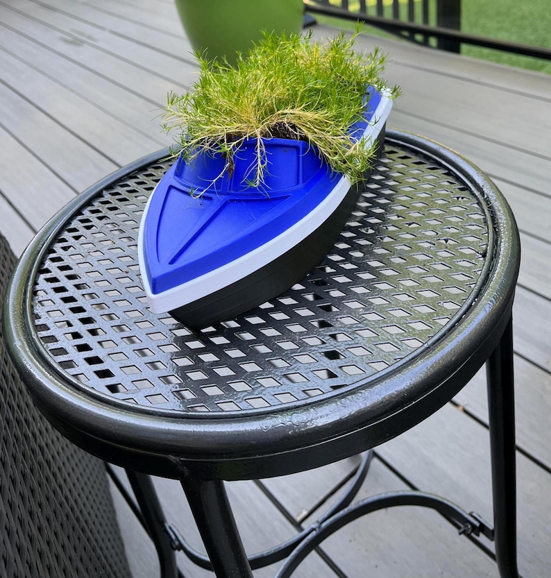 May include: A blue, white, and black boat-shaped planter filled with green plants. The planter sits on a black metal table with a patterned surface. The table has a round top and three legs. The background includes a green pot and wooden decking.
