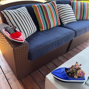 May include: A navy blue outdoor sofa with striped and solid throw cushions. A red boat-shaped tray holds remote controls. A blue boat-shaped planter with flowers sits on a concrete table. Hanging flower baskets are in the background.