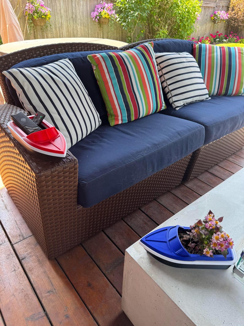 May include: A navy blue outdoor sofa with striped and solid throw cushions. A red boat-shaped tray holds remote controls. A blue boat-shaped planter with flowers sits on a concrete table. Hanging flower baskets are in the background.