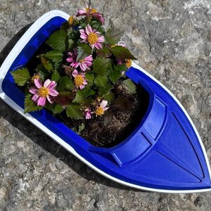May include: A blue boat-shaped planter with a white trim, filled with green foliage and pink flowers. The planter sits on a textured grey stone surface, creating a contrast between the vibrant colours of the flowers and the planter.
