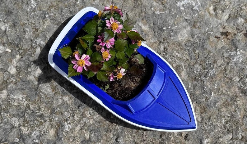 May include: A blue boat-shaped planter with a white trim, filled with green foliage and pink flowers. The planter sits on a textured grey stone surface, creating a contrast between the vibrant colours of the flowers and the planter.