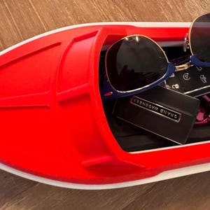 May include: A red and white boat-shaped tray containing sunglasses, keys, and a small black box. The sunglasses have blue frames. The tray sits on a wooden surface.