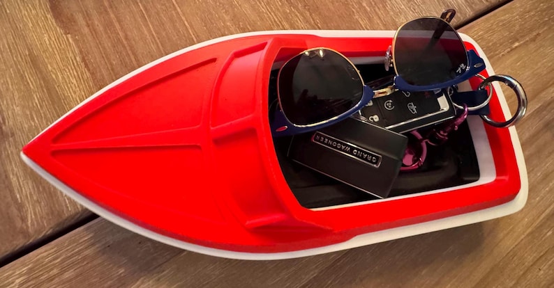 May include: A red and white boat-shaped tray containing sunglasses, keys, and a small black box. The sunglasses have blue frames. The tray sits on a wooden surface.