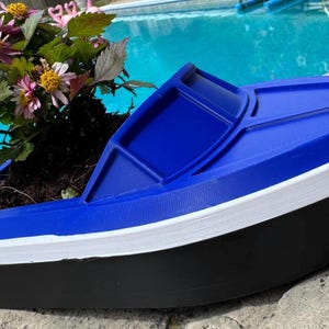 May include: A boat-shaped planter in blue, white, and black, containing soil and flowering plants. The planter features a blue top, white trim, and a black base. The flowers are pink and yellow.