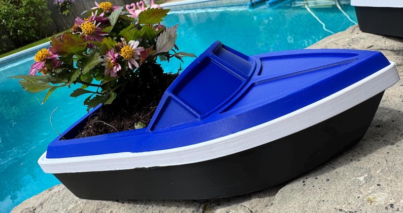May include: A boat-shaped planter in blue, white, and black, containing soil and flowering plants. The planter features a blue top, white trim, and a black base. The flowers are pink and yellow.