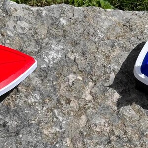 May include: Two boat-shaped planters, one red and one blue, are on a stone surface. The red planter is empty, while the blue one contains soil and flowering plants with pink and yellow blooms. Both have white trim.