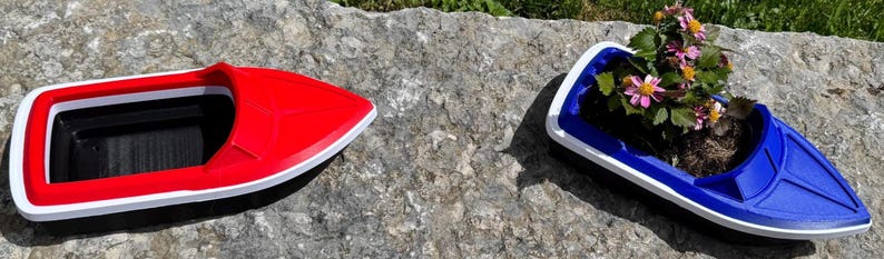 May include: Two boat-shaped planters, one red and one blue, are on a stone surface. The red planter is empty, while the blue one contains soil and flowering plants with pink and yellow blooms. Both have white trim.