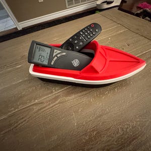May include: A red plastic boat-shaped organiser holding two black remote controls. The organiser has a white trim and a black base. One remote displays a digital temperature reading.