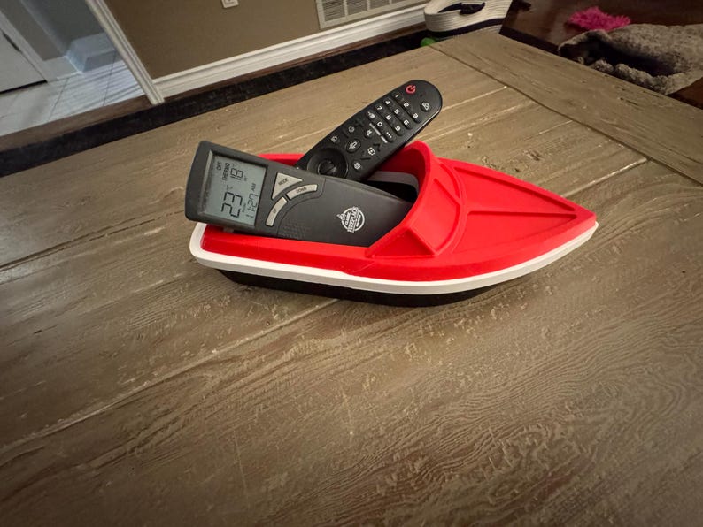 May include: A red plastic boat-shaped organiser holding two black remote controls. The organiser has a white trim and a black base. One remote displays a digital temperature reading.