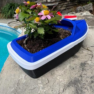 May include: A boat-shaped planter in blue, white, and black, containing soil and flowering plants. The planter sits on a stone surface beside a pool. The flowers have pink and yellow petals.