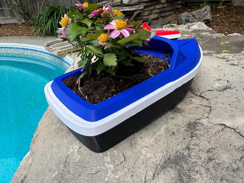 May include: A boat-shaped planter in blue, white, and black, containing soil and flowering plants. The planter sits on a stone surface beside a pool. The flowers have pink and yellow petals.