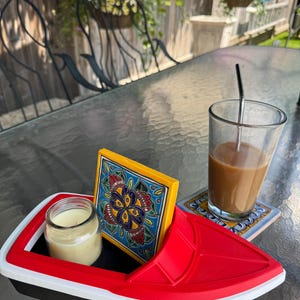May include: A red, white, and black boat-shaped tray holds a small candle in a glass jar and a decorative tile with a floral design. A glass of coffee with a straw and a coaster are also on the table.