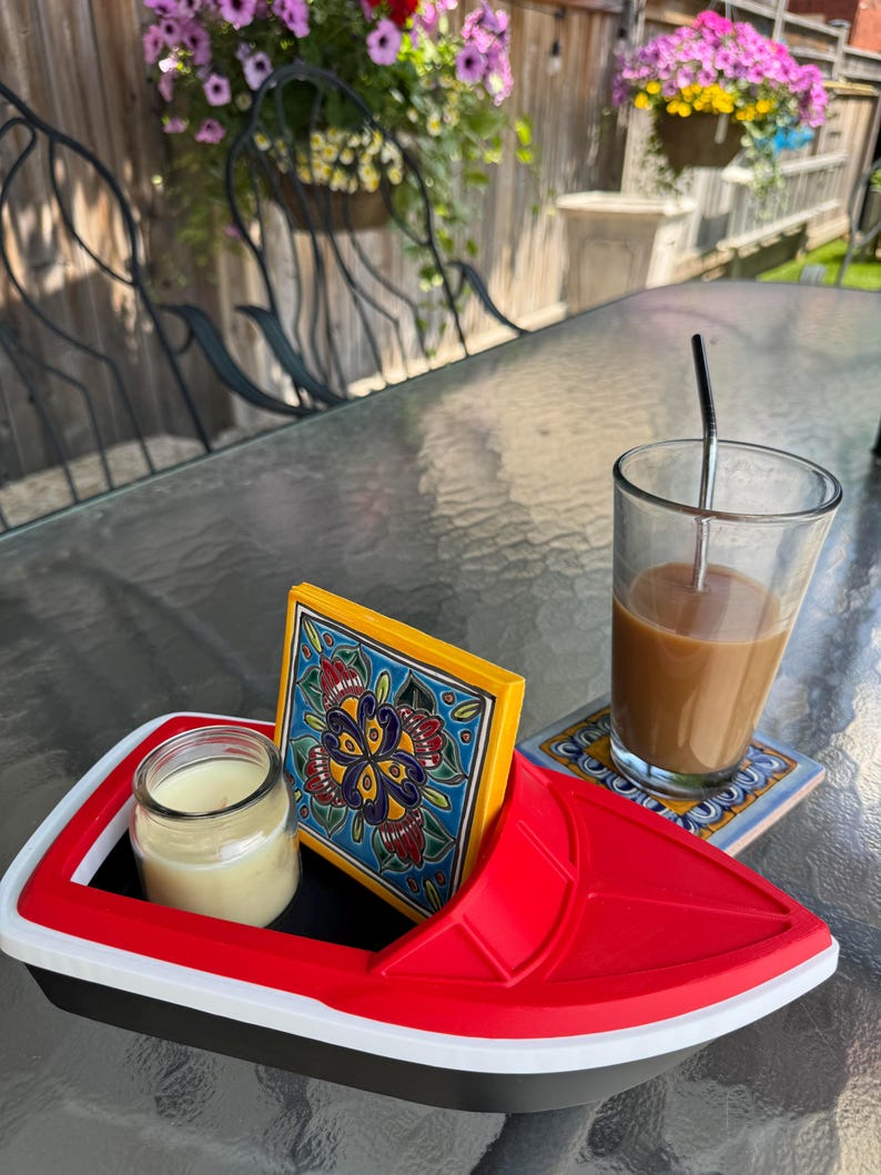 May include: A red, white, and black boat-shaped tray holds a small candle in a glass jar and a decorative tile with a floral design. A glass of coffee with a straw and a coaster are also on the table.