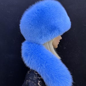May include: A bright blue fur hat and scarf combination. The hat has a rounded crown, and the scarf falls over the shoulders. The fur looks soft and fluffy, with a uniform colour. The background is a plain black.