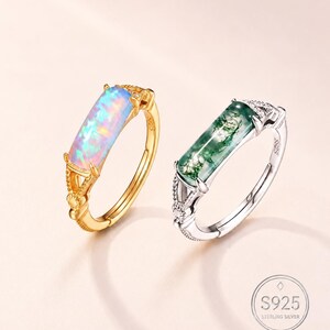 May include: Two rings featuring rectangular stones set in metal bands. The left ring is gold-toned with an iridescent opal stone. The right ring is silver-toned with a green moss agate stone. Both bands are marked S925 sterling silver.