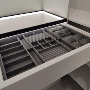 Può includere: Un organizzatore di gioielli bianco con più scomparti rivestiti in velluto grigio. Gli scomparti sono progettati per contenere anelli, bracciali, collane e orologi. L'organizzatore è integrato in un mobile bianco con illuminazione a incasso.
