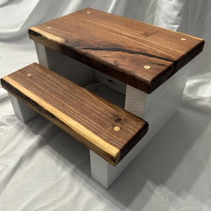 May include: A two-step wooden stool with a natural wood top and a white painted base. The top step is 12 inches long and the bottom step is 10 inches long. The wood has a natural grain and is secured with small wooden pegs.